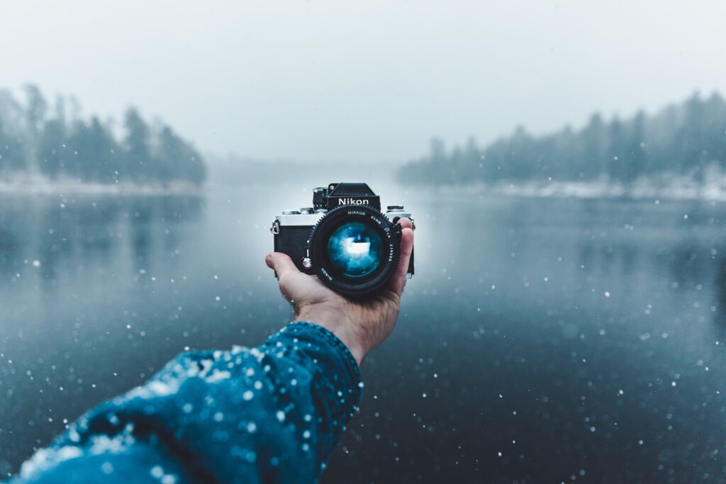 Someone holding a Nikon camera facing the camera while extended in front of a lake surrounded by forest.